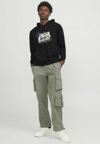 Sudadera negra con capucha y estampado gráfico, combinada con pantalones cargo verde claro que cuentan con múltiples bolsillos, complementados con zapatillas blancas.