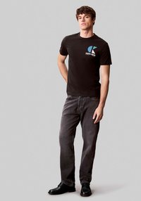 Camiseta negra de algodón con un logo azul y blanco en el pecho, combinada con jeans gris oscuro y zapatos negros, posando contra un fondo gris claro.