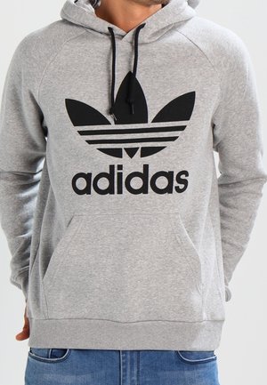 Mężczyzna ubrany w jasnoszary kapturowy sweter Adidas z czarnym logo trefoil i napisem, ręce za plecami, w połączeniu z niebieskimi jeansami.