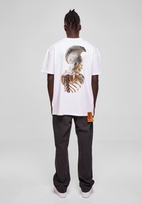Hvit T-skjorte med et detaljert grafisk bilde av en hjelmiført figur på baksiden, kombinert med mørke jeans og et oransje merke.