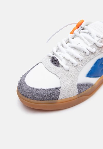Λευκό αθλητικό παπούτσι με γκρι σουέντ μύτη, μπλε λογότυπο στη πλευρά, λευκά κορδόνια, πορτοκαλί κλείδωμα κορδονιών και καουτσούκ σόλα σε λευκό φόντο.
