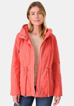 Korallfarbene, leichte Jacke mit Kapuze, Kordelzug in der Taille, metallischen Akzenten und Reißverschlüssen. Kombiniert mit einem strukturierten beigen Pullover darunter.