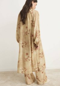 Lungo vestito fluido realizzato in tessuto beige con motivi floreali marroni. Presenta maniche lunghe e una fessura sulla schiena per facilità di movimento.