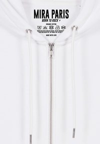 Felpa bianca con zip realizzata in cotone biologico, dotata di una zip in metallo argentato, cordini regolabili e un'etichetta stampata con le istruzioni per la cura.
