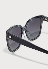 Ochelari de soare cu rame pătrate, dintr-un material gri transparent, cu un model houndstooth. Brațele negre au un accent auriu care prezintă detalii cu logo.