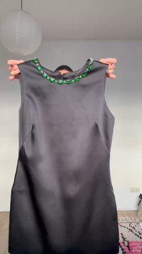 Abito nero senza maniche sollevato, con dettagli di gemme verde smeraldo intorno allo scollo, su uno sfondo interno semplice.