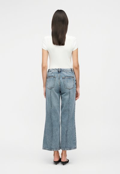 Γυναίκα που στέκεται με την πλάτη, φοράει λευκό κοντομάνικο τοπ, μπλε τζιν με φαρδιά πόδια και μαύρα σανδάλια με τακούνι σε λευκό φόντο.