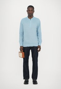 Lyseblå henley skjorte med tre knappelukking, kombinert med mørk denimbukser. Modellen holder en brun firkantet håndveske.