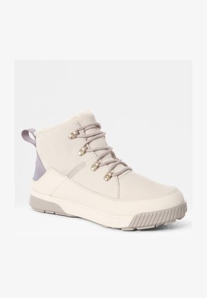 Bota alta beige con acentos grises, cuello de pelaje sintético texturizado, cierre con cordones y ojales metálicos, y suela resistente para tracción.