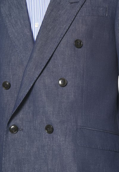 Sötétkék, kétsoros blazer, texturált anyagból, fekete gombokkal és egyetlen mellzsebbel. Csíkozott ing látható alatta.