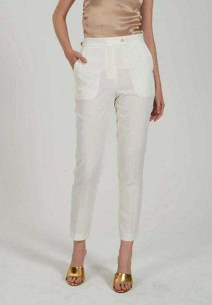 Pantaloni bianchi a taglio slim con vita alta e tasche frontali, abbinati a tacchi open-toe dorati metallizzati e una top beige senza maniche.