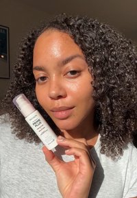 Revlon Prime Plus Perfecting Smoothing Primer en una botella blanca opaca con un dispensador rosa, sostenida por una persona con cabello rizado y piel clara.