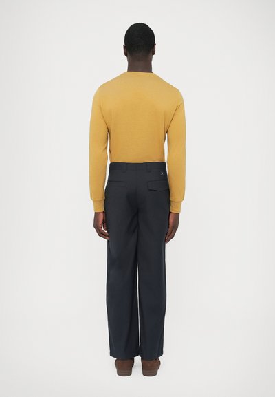 Maglione color senape a maniche lunghe abbinato a pantaloni blu navy a gamba larga, con tasche posteriori e una trama liscia. Scarpe marroni completano l'insieme.