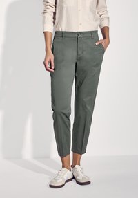 Grüne Baumwollhosen im Slim-Fit, mit seitlichen Taschen und Knopfverschluss, kombiniert mit weißen Sneakers und einer hellbeigen Bluse.