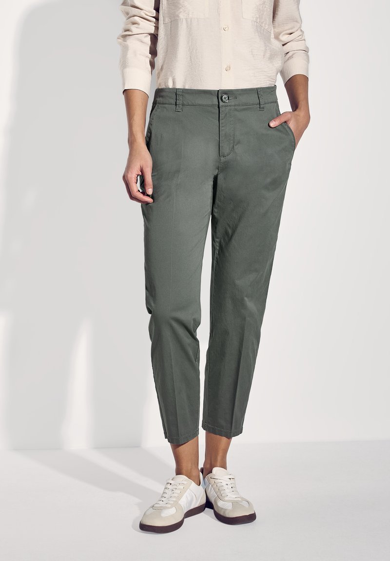 Grüne Baumwollhosen im Slim-Fit, mit seitlichen Taschen und Knopfverschluss, kombiniert mit weißen Sneakers und einer hellbeigen Bluse.