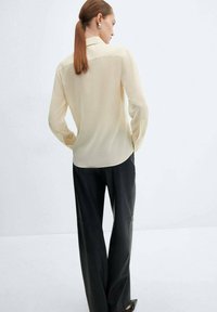 Donna con coda di cavallo castana che indossa una blusa a maniche lunghe crema e pantaloni neri a gamba larga, in piedi con le mani nelle tasche, rivolta verso destra.
