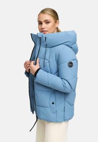 Ljust blå uppblåst jacka med huva, dragkedja framtill, quiltat mönster, sidofickor och en logotyp på vänster arm. Strukturerat, syntetiskt material.