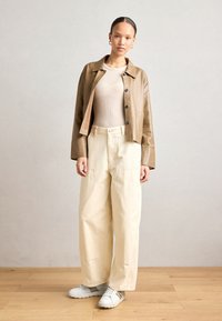 Giubbotto in pelle marrone corto, top beige a coste, pantaloni ampi crema, sneakers bianche, con dettagli a bottoni e vestibilità oversize.
