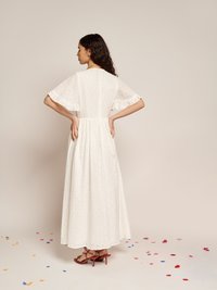 Vestido maxi branco com padrão de ilhós, mangas curtas com volantes, cintura ajustada e saia fluida. Usado com sandálias de salto alto vermelhas.