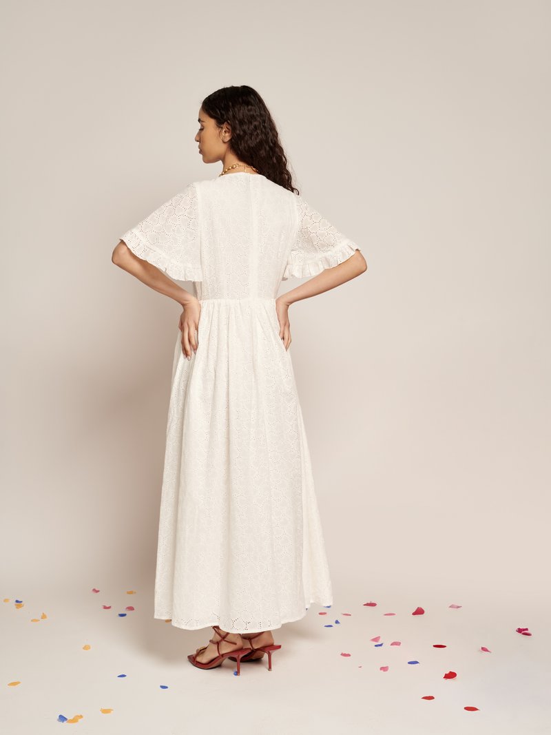 Vestido maxi branco com padrão de ilhós, mangas curtas com volantes, cintura ajustada e saia fluida. Usado com sandálias de salto alto vermelhas.