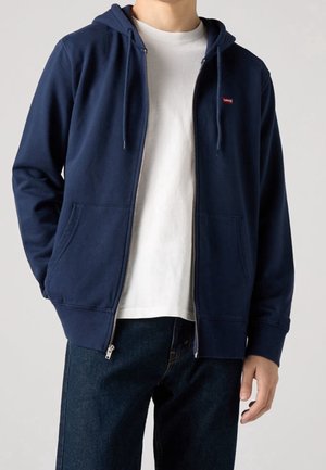 Man wearing navy zip-up hoodie with front pockets over white t-shirt and dark blue jeans against plain background.