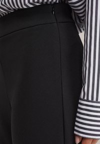 Črne krojene hlače se ponašajo s strukturirano silhueto in stranskim zadrgo, v kombinaciji z majico z našitki z lepenko za kontrast.