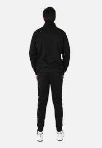 Tuta nera con colletto alto, polsini a coste e vita elastica. Il modello indossa sneakers bianche e verdi. Vista posteriore mostrata.