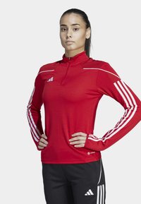Maglietta sportive rossa a maniche lunghe con colletto con zip, caratterizzata da accenti bianchi a tre strisce sulle maniche e logo sul petto.
