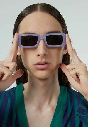 Paarse rechthoekige zonnebril met donkere lenzen, vastgehouden bij de slapen, gedragen door een persoon in een groen en blauw gestreept shirt tegen een effen achtergrond.