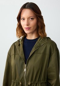 Giacca a zip verde oliva con cappuccio, dotata di vita regolabile con cordoncino, zip in metallo e texture liscia, abbinata a una maglia blu navy sotto.