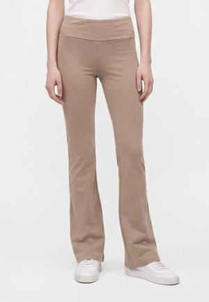 Personne portant un pantalon évasé beige, des baskets blanches et un haut blanc, debout devant un fond clair uni.