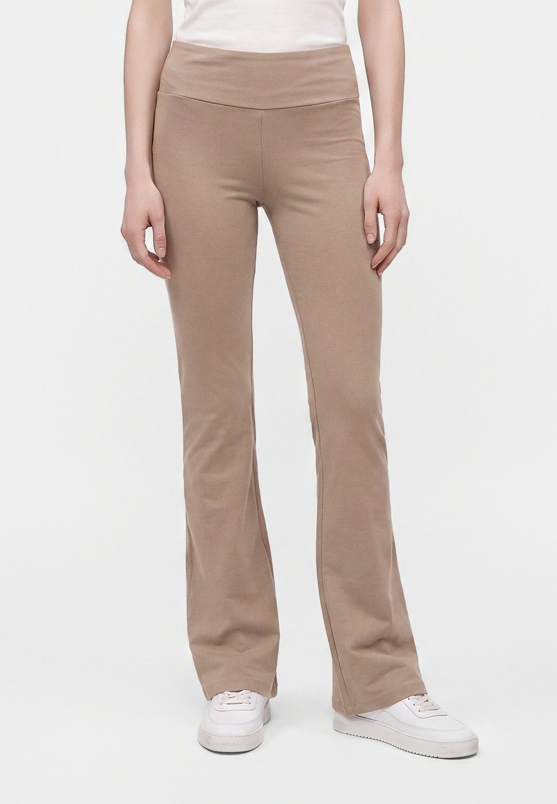 Personne portant un pantalon évasé beige, des baskets blanches et un haut blanc, debout devant un fond clair uni.