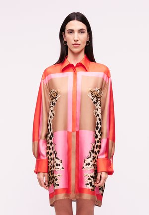 Mujer con cabello largo y oscuro que lleva un vestido camisa suelto de manga larga con bloques de color rojo, rosa y beige, y estampado de leopardo espejado en la parte delantera.