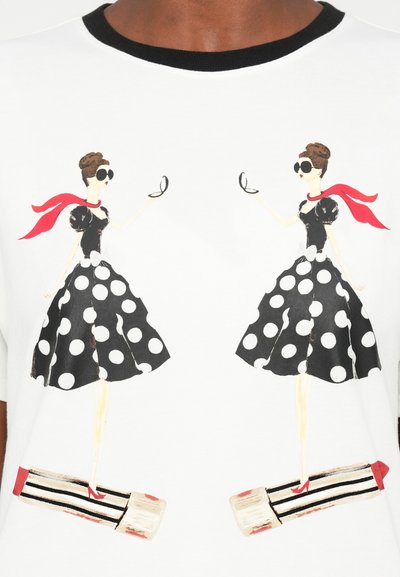Vitt t-shirt med två stiliserade illustrationer av kvinnor i svarta prickiga klänningar, röda detaljer och stora solglasögon, som håller speglar.
