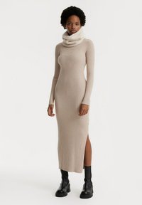 Beige geribbelde gebreide jurk met een hoge kraag, strakke silhouet, zijsplit en gecombineerd met zwarte enkellaarsjes.