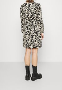 Kvinna i en svart klänning med vit blommig tryck, knuten i midjan, parat med svarta, tjocka stövlar som når till vaden, står med ryggen vänd.