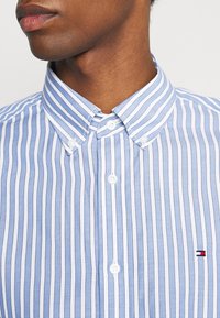 Lichtblauw-wit gestreept overhemd met knopen en een puntige kraag, voorzien van een klein logo met rode en blauwe accenten. Gladde stoftextuur.