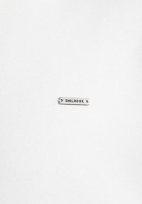 Malá obdélníková stříbrná kovová štítka s vyraženým slovem "SMILODOX", umístěná na obyčejném bílém textilu.