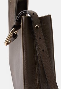 Καφέ υφασμάτινη δερμάτινη τσάντα με δομημένο σχέδιο, διαθέτοντας μακρύ λουρί και χρυσές μεταλλικές λεπτομέρειες για κλείσιμο και ρύθμιση.