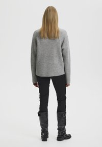 Suéter gris con un tejido texturizado, corte holgado. Pantalones negros y botas negras hasta la rodilla con hebillas. Vista trasera, fondo blanco liso.