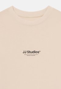 T-shirt beige à manches courtes avec col rond, arborant le logo en texte noir "JJ Studios" et des détails supplémentaires en dessous. Texture en coton doux.