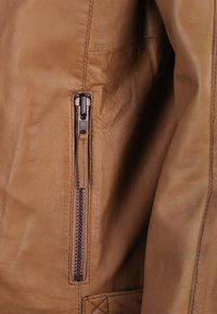 Braune Lederjacke mit einer Seitentasche mit YKK-Reißverschluss, samtiger Textur und dezenten Nähdetails.