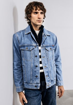 Ung mand med krøllet brunt hår iført en lyseblå denimjakke over en sort-hvid stribet sweater, stående mod en hvid væg.