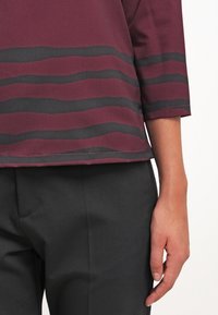 Bordeauxrote Bluse mit horizontalen schwarzen Streifen, 3/4-Ärmel, glatte Textur; kombiniert mit schwarzen Hosen. Fokus auf der oberen Hälfte mit sichtbarer Hand.