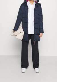 Marineblå puffet jakke med hætte, der har et horisontalt quiltet design og en rødstribet inderfor. Modellen holder en beige tæppe.