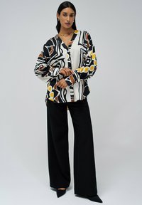 Blouse à manches longues présentant un motif abstrait audacieux en noir, blanc et jaune, assortie à un pantalon noir à jambes larges et des escarpins à bout pointu.