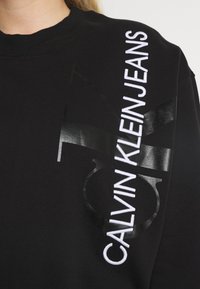 Sudadera negra con tejido texturizado, un gran logotipo en negro brillante y texto bordado en blanco a lo largo de la manga izquierda.