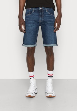 Hombre con shorts de mezclilla azul oscuro con dobladillo, zapatillas blancas de caña alta y calcetines blancos con rayas rojas y negras.