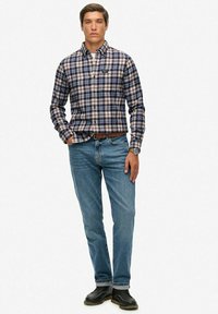 Herren-Karobluse in Marineblau, Weiß und Beige; kombiniert mit hellblauen Jeans und schwarzen Schuhen. Glatte Textur, klassischer Schnitt und zwei Brusttaschen.