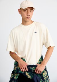 Kremo spalvos per didelė t-shirt su trumpomis rankovėmis, papuošta mažu tamsiai mėlynu logotipu. Modelis laiko daugiaskarę pūkuotą striukę su žaliais ir mėlynais akcentais.